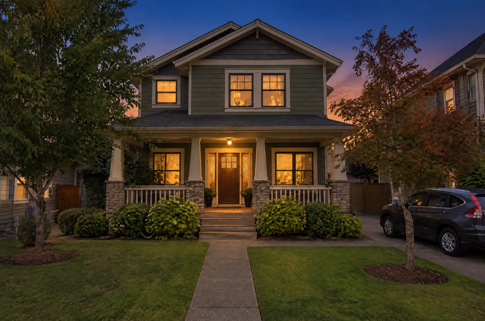 The same house at dusk: blue sky, warm light in its windows and the porch light on.